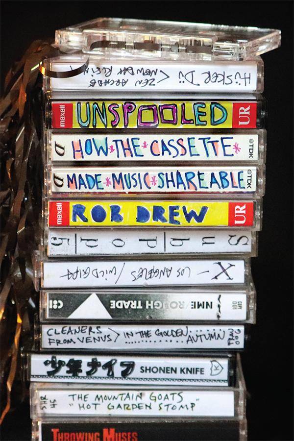 Cover of Unspooled: How the Cassette Made Music Shareables by Rob Drew. Cover features a photograph of a stack of cassette tapes in their cases against a black background. Cassette titles, featured on the side of the cases, include Throwing Muses, The Mountain Goats "Hot Garden Stomp," Cleaners from Venus In the Golden Autumn, NME/Rough Trade, Los Angeles/Wild Gift, SubPop, and Husker Du Zen Arcade New Day Rising.